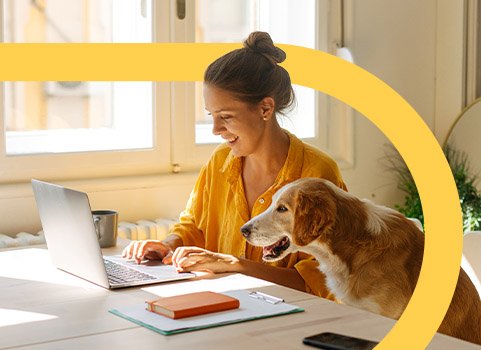 Woman in yellow shirt using laptop with dog beside her.