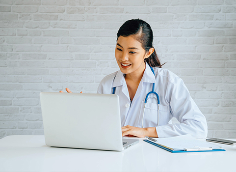 A woman with medium dark skin and dark hair wears a lab coat and a stethoscope and works on a computer