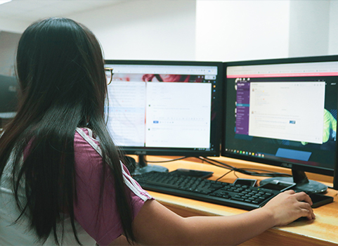 A woman with medium dark skin and dark hair watches two computer monitors 