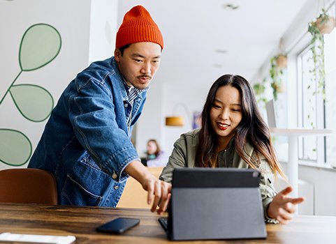 A woman with dark hair sits in front of a tablet, talking to a man in a red beanie who leans over her, pointing at the tablet