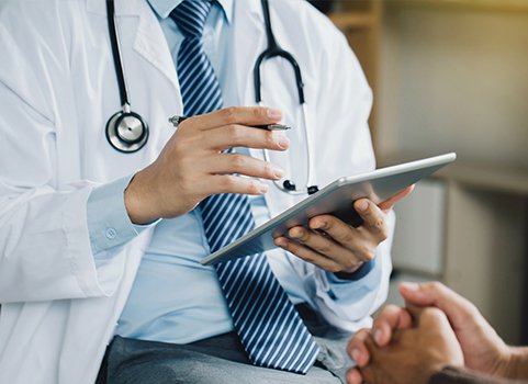 A person in a suit and tie wearing a lab coat and a stethoscope, holding an ipad and gesturing to another person with folded hands