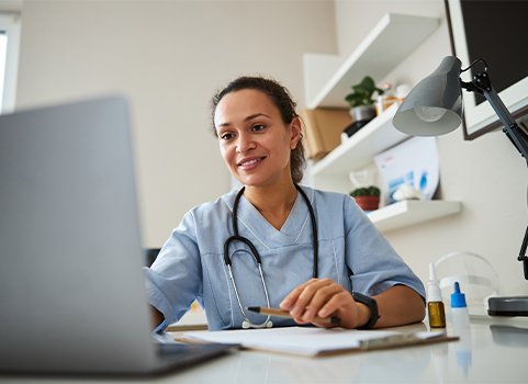 A woman wearing scrubs and a stethoscope and working on a laptop 